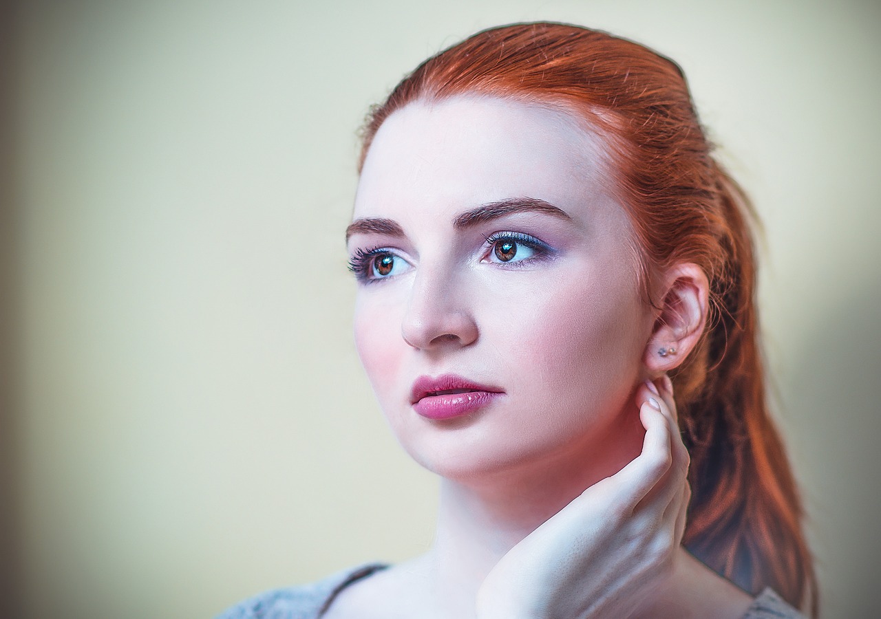 Portret rudowłosej kobiety o jasnej cerze i niebieskich oczach, z dłonią przy twarzy i delikatną biżuterią w uszach.